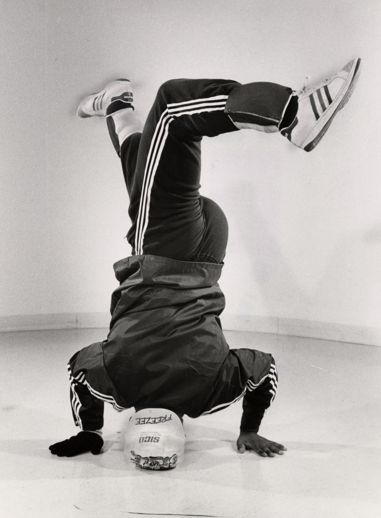 Whole lotta breakin' going on, by Tony Brock, 1984. Exhibit caption reads, "Nigel 'Sugar Pop' Walters, an 18 year-old student at North York's Earl Haig Collegiate, shows off his scissor kick during a headstand. A few days after this photo was taken, 'Breakdance '84,' Toronto's 'first ever high school street dance show,' was held at Riverdale Collegiate." Image from Toronto Public Library under Toronto Star License.